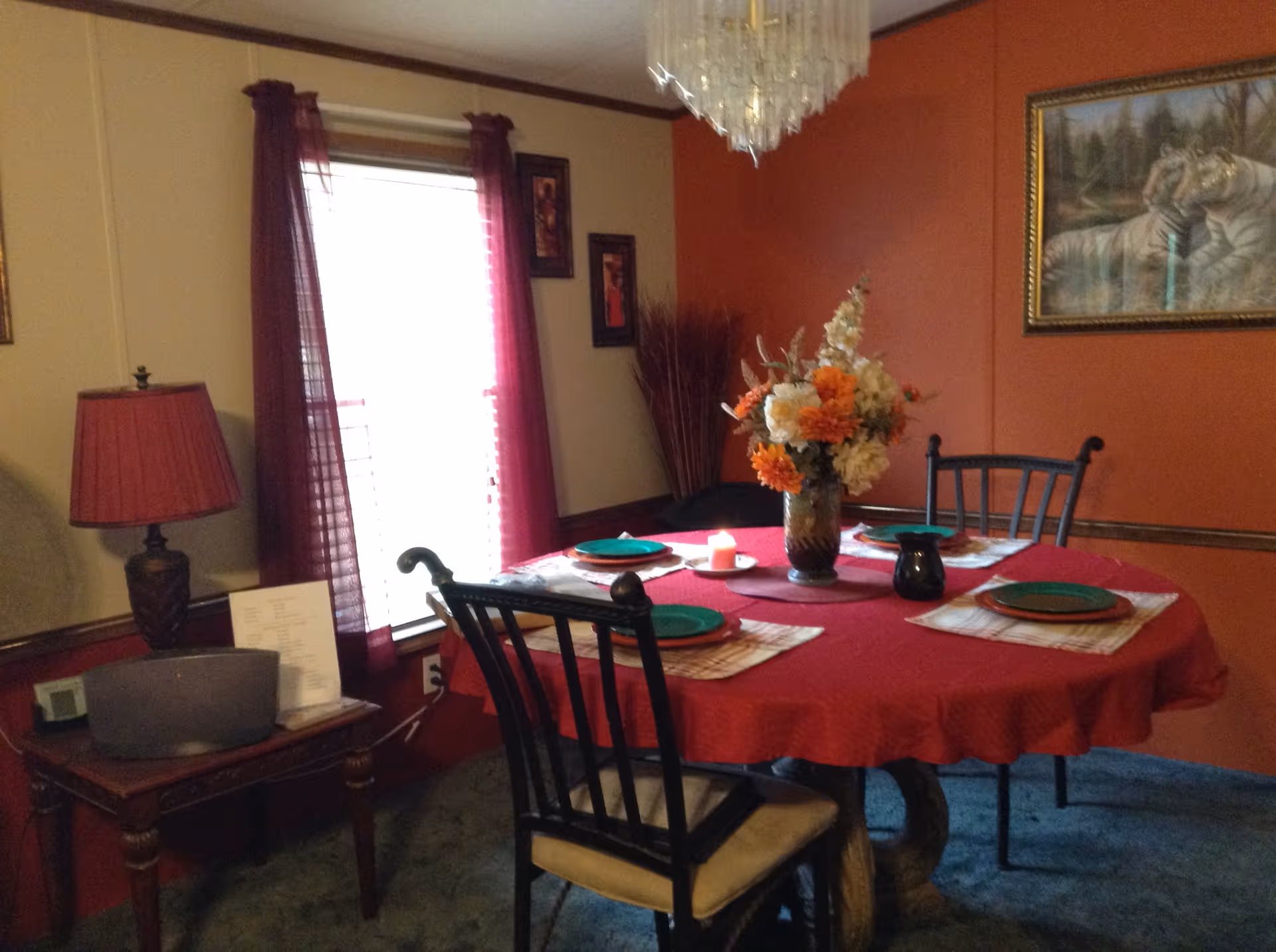 A dining room with a round table covered with a red tablecloth, set with four place settings including green plates and placemats. A vase with orange and white flowers is in the center of the table, along with a small candle and a black pitcher. The room has orange and beige walls, a window with red curtains, a side table with a lamp and a speaker, and a framed painting of two white tigers on the wall.