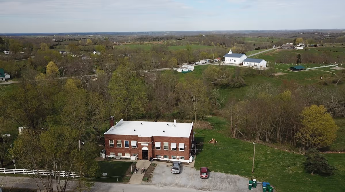 Aerial view of a brick two-story building with a white roof set in a rural landscape of trees and fields.