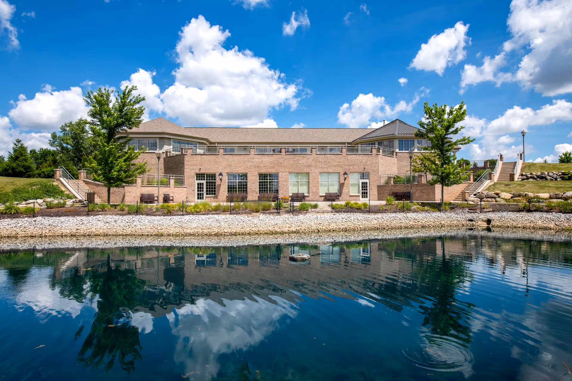 A brick senior living facility sits along a pond with trees and a blue sky.