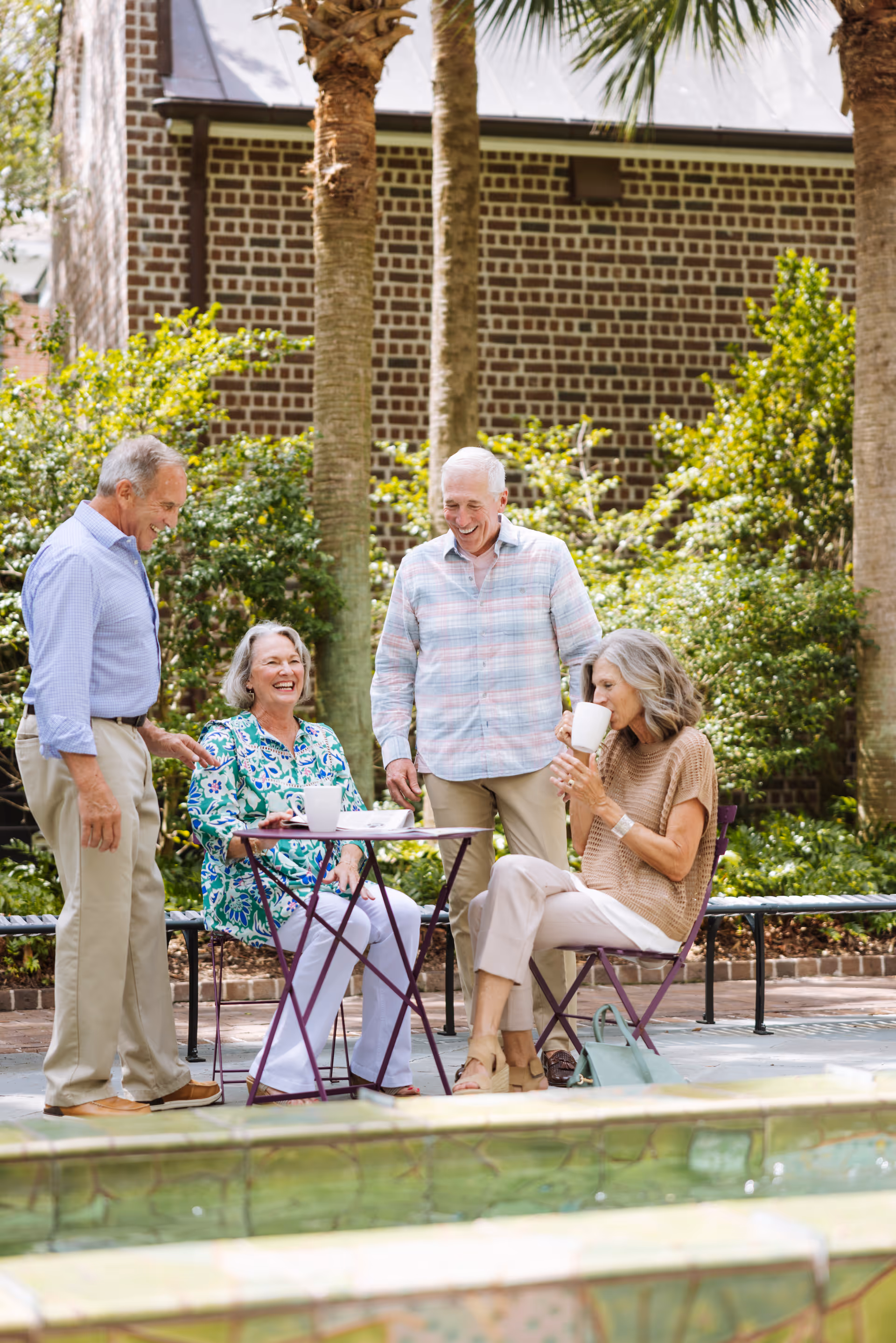 Four seniors chat and sip coffee at a sunny outdoor courtyard near a brick building.