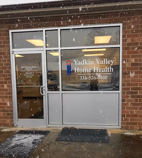 Brick building entrance with glass doors displaying Yadkin Valley Home Health signage in snow.