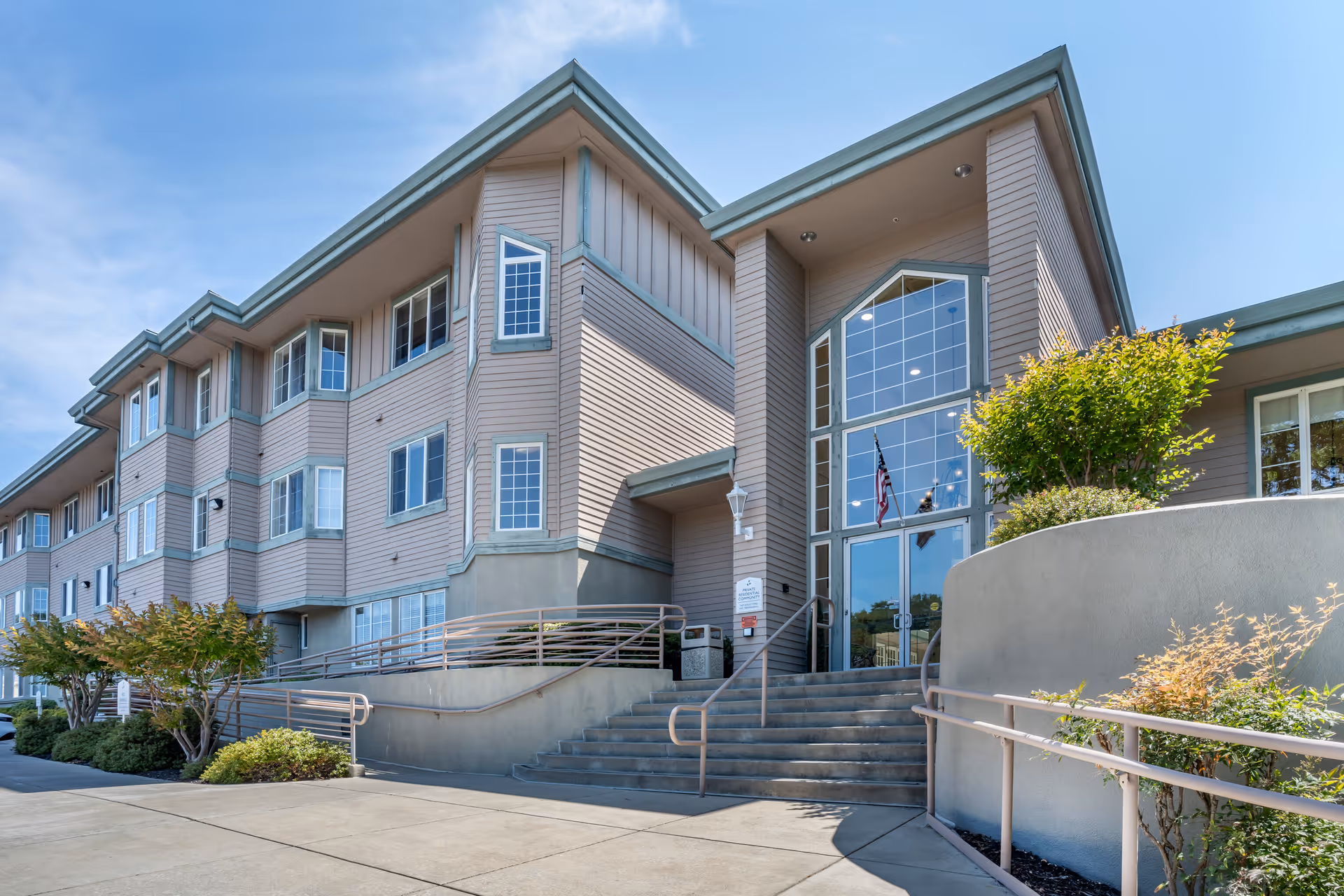 Entrance and facade of a multi-story senior living building with stairs, a wheelchair ramp, large glass entryway, and landscaping.