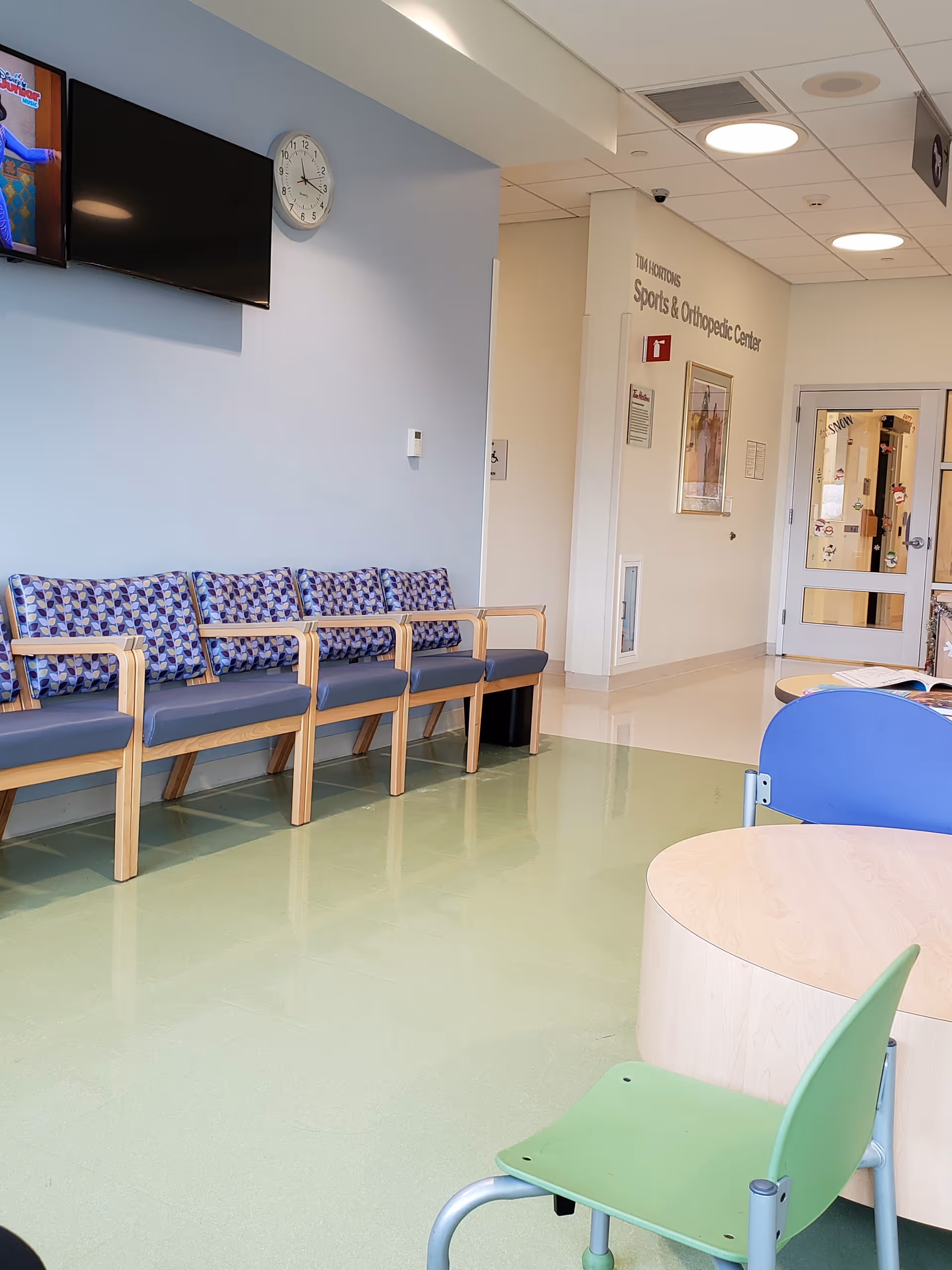 Clinic waiting area with blue cushioned chairs, a wall-mounted TV, and a clock.