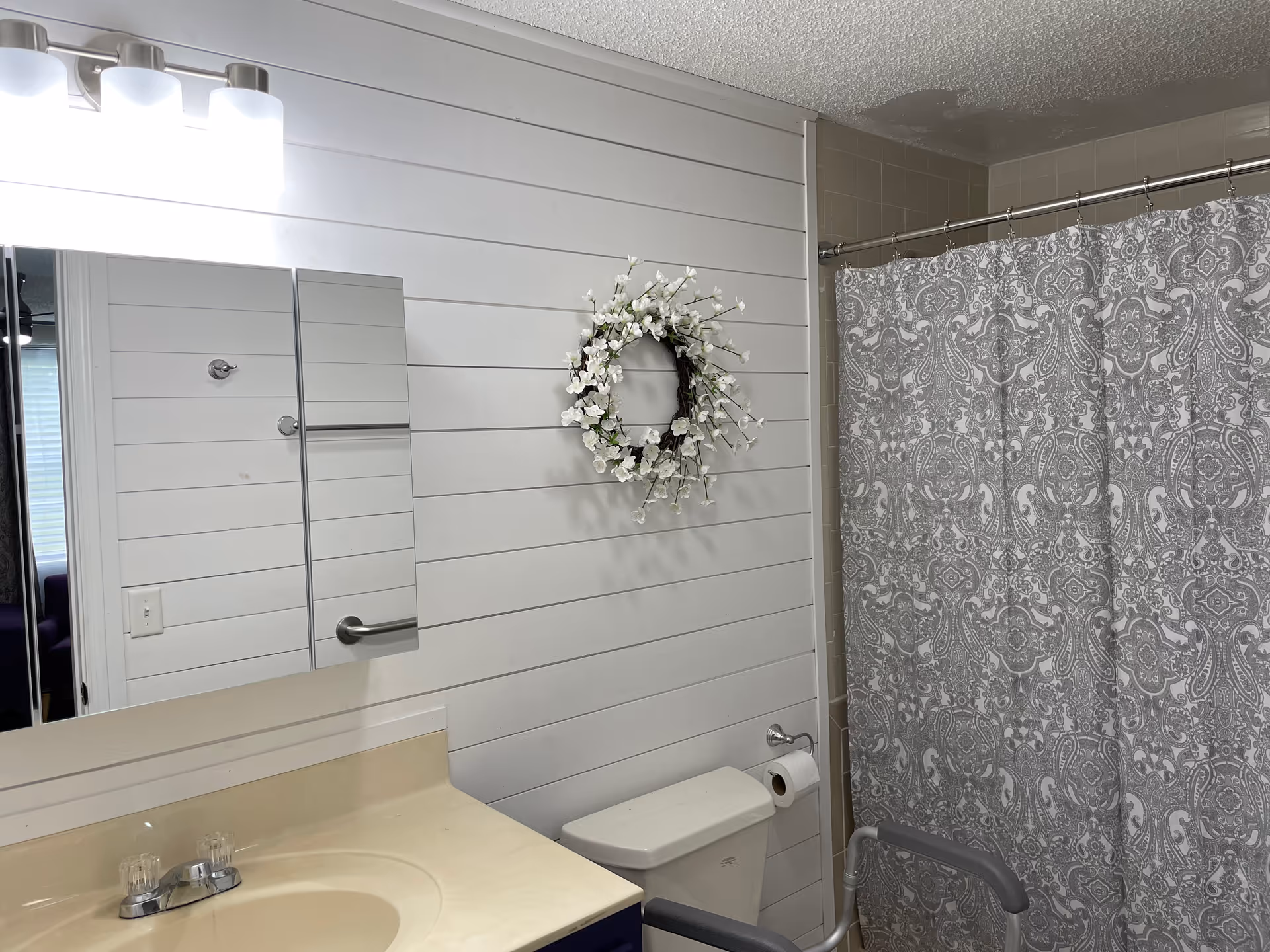 Bathroom with white shiplap walls, sink, mirror, wreath, toilet, and patterned shower curtain.