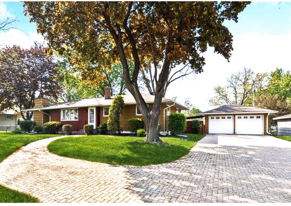 Front view of a single-story house with curved brick driveway, trees, and a green lawn.