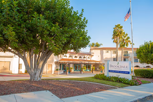 Front entrance of the Brookdale San Marcos senior living facility with a sign, flagpole, and trees in front of a Mediterranean-style building.