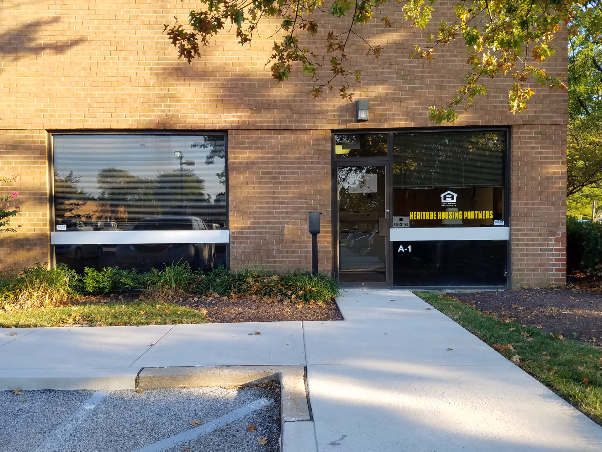Brick building entrance with glass doors and large front windows, shaded by trees.