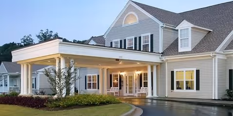 Exterior view of The Willows of Easley senior living facility at dusk, showing a large, two-story building with beige siding, white trim, black shutters, and a covered entrance with columns. The entrance is illuminated with warm lights, and there is a curved driveway and landscaped greenery in front.