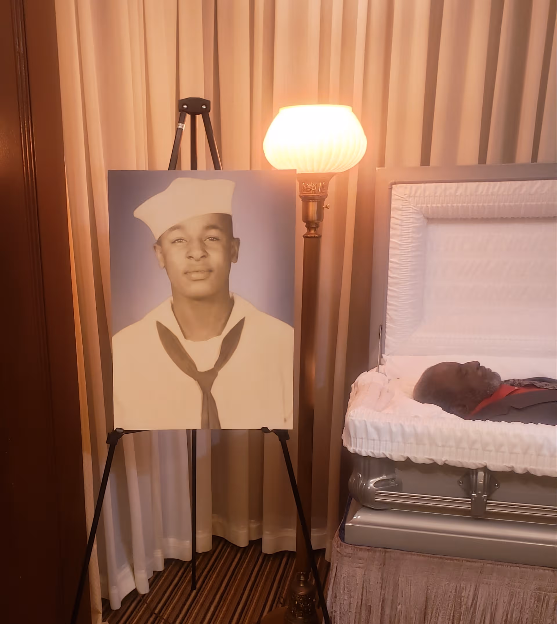 Portrait of a man in a sailor uniform beside an open casket in a lit room.