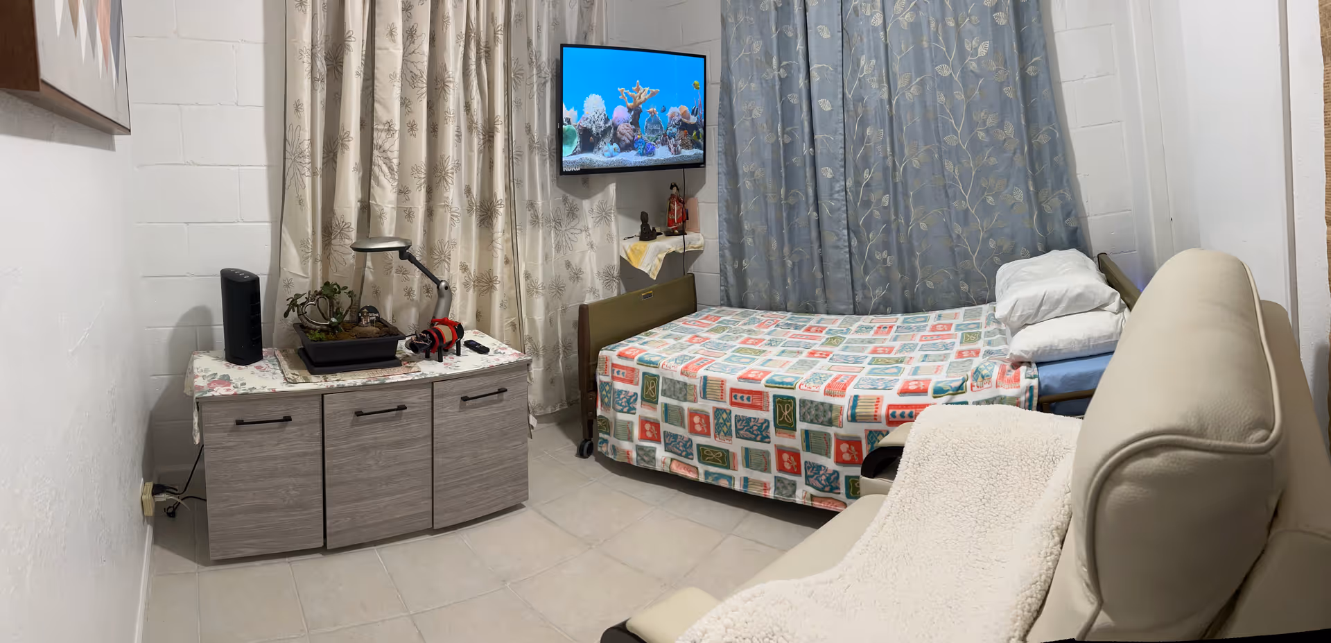 Compact private bedroom with bed, dresser, TV, and reclining chair in a senior facility.