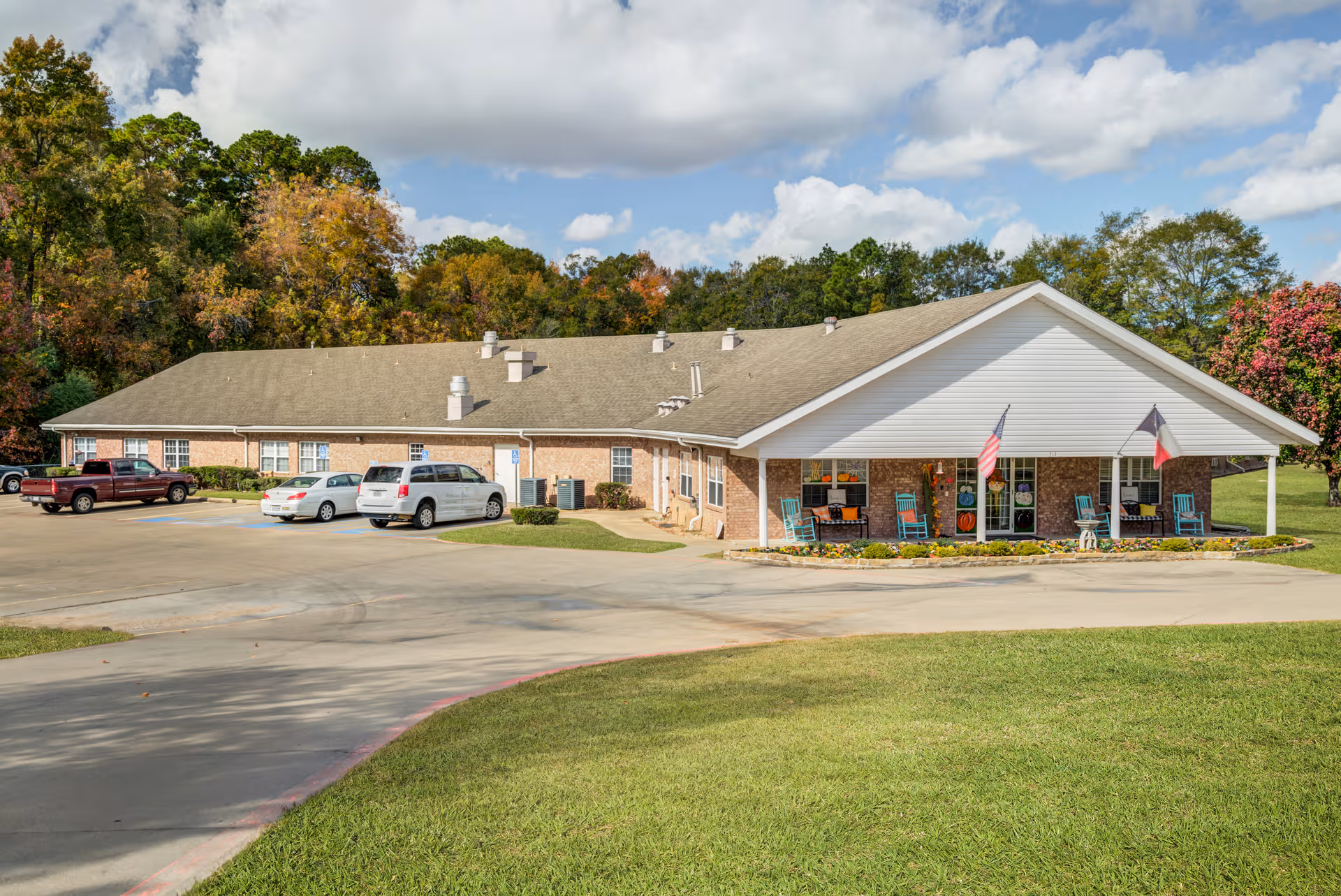 Single-story brick senior living facility with a covered front entrance, parked cars, and green lawn.