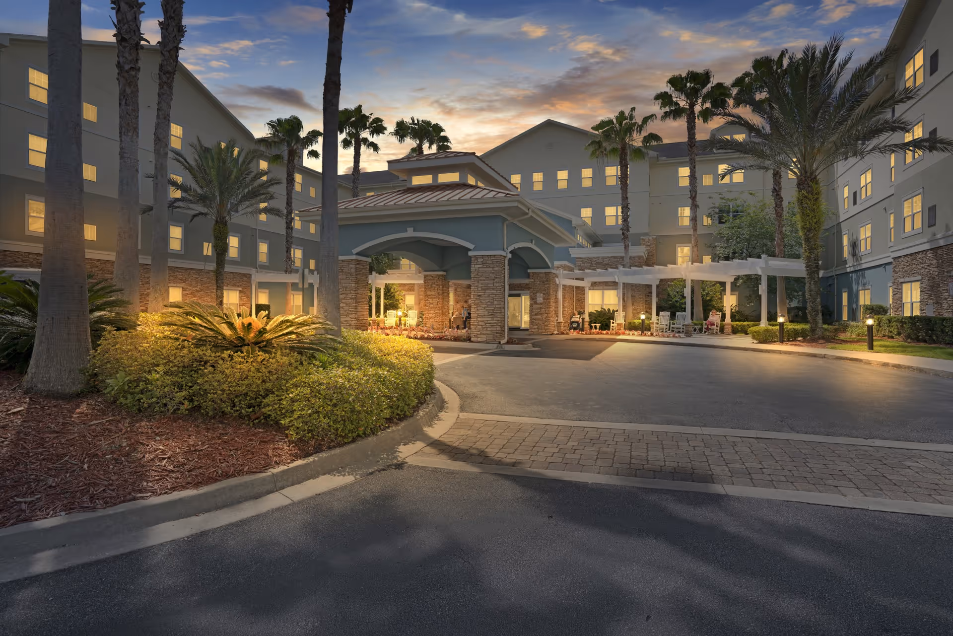Exterior view of Camellia At Deerwood senior living facility at dusk, showing a large building with multiple windows lit from inside, palm trees, landscaped bushes, and a covered entrance with outdoor seating under a pergola.