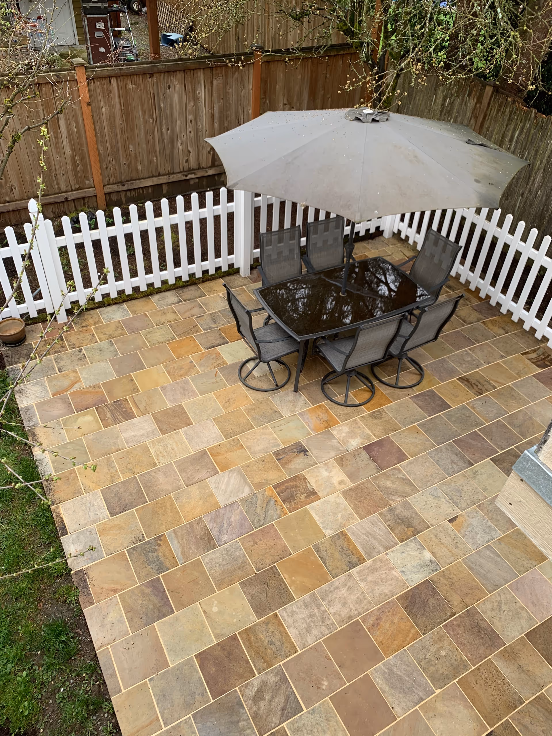 Outdoor tiled patio with glass table, chairs, and umbrella in a fenced yard.