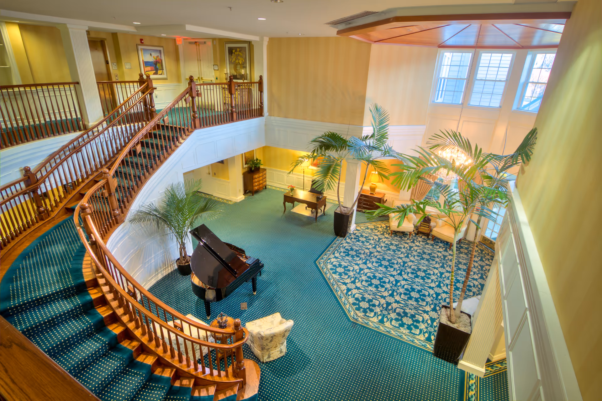 Interior view of a senior living facility lobby with a curved wooden staircase, green carpet with white dots, a black grand piano, potted palm plants, a seating area with two armchairs on a patterned blue rug, and large windows letting in natural light.