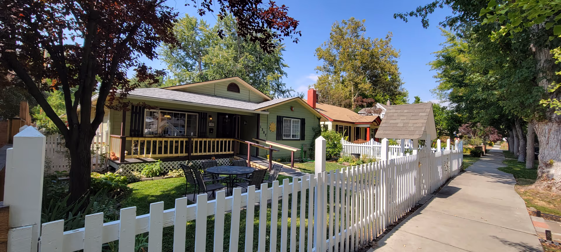 Green single-story Ivy Place Residence behind a white picket fence with outdoor seating.