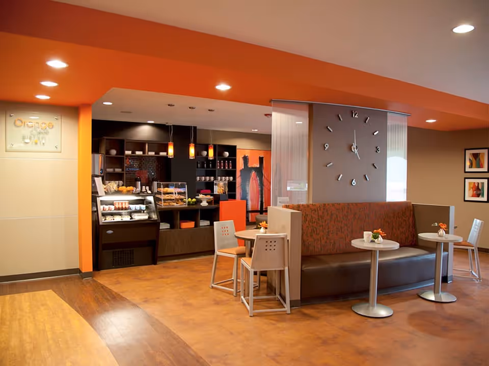 Interior view of a cozy cafe area with orange accents, featuring a small counter with baked goods and beverages, modern pendant lights, a large wall clock, and seating with tables and chairs arranged for casual dining.