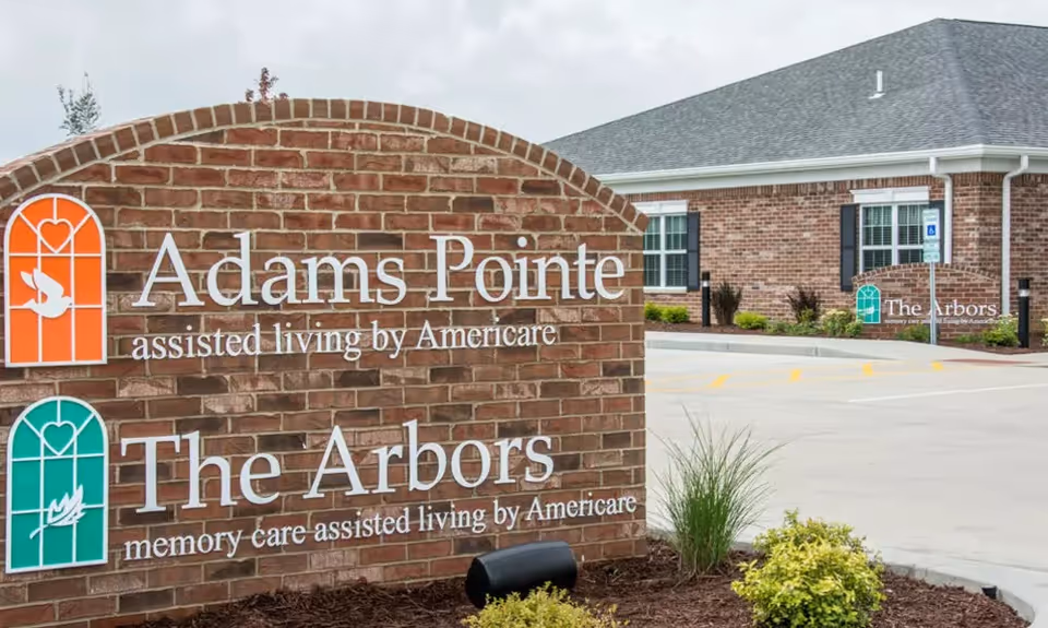 A brick sign at the entrance of a senior living facility displaying the names Adams Pointe assisted living by Americare and The Arbors memory care assisted living by Americare. In the background, there is a brick building with windows and a parking lot.