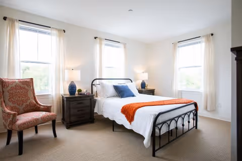 A bright bedroom with three large windows covered by light curtains. The room features a black metal bed frame with white bedding, a blue pillow, and an orange throw blanket. There are two dark wooden nightstands on either side of the bed, each with a blue lamp and decorative items. A patterned upholstered armchair is placed near one window.