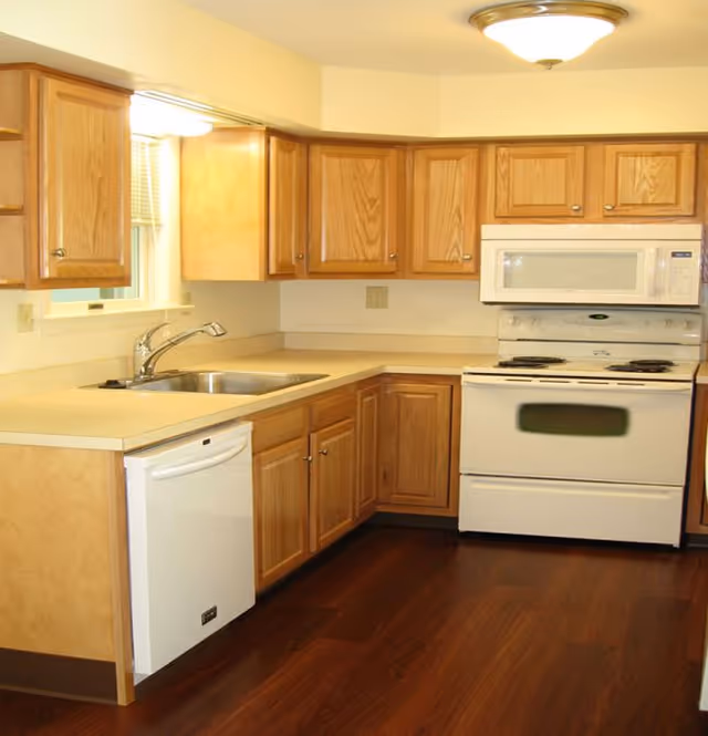 A clean kitchen with wooden cabinets, a white dishwasher, a stainless steel sink, a white electric stove with an oven, a white microwave above the stove, and dark wood flooring.