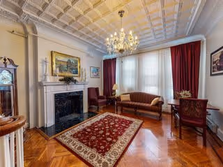 A cozy living room with a chandelier hanging from a decorative ceiling. The room features a fireplace with a painting above it, a red patterned rug on a wooden floor, a sofa, armchairs, a small table with a lamp, and large windows with red curtains.
