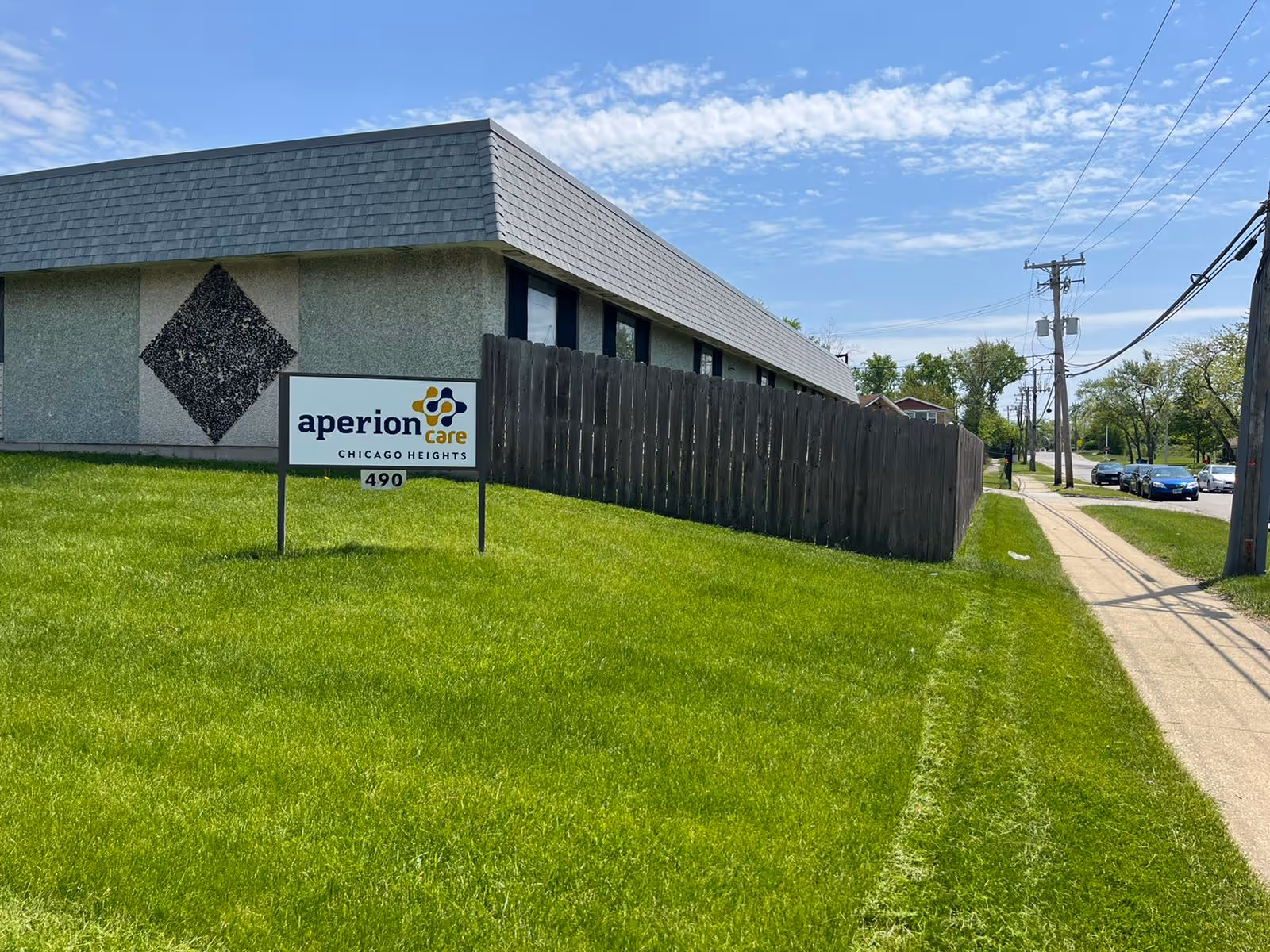 Exterior view of Aperion Care Chicago Heights building with a green lawn and a sign displaying the facility name and address number 490. A wooden fence runs alongside the building, and a sidewalk with utility poles and cars is visible on the right side under a partly cloudy sky.