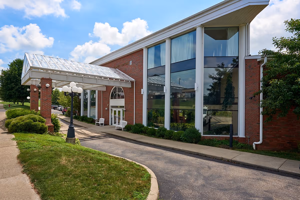 Exterior view of the Country Club Retirement Campus building entrance with a covered driveway, large windows, brick walls, and a sidewalk surrounded by green bushes and trees under a partly cloudy blue sky.