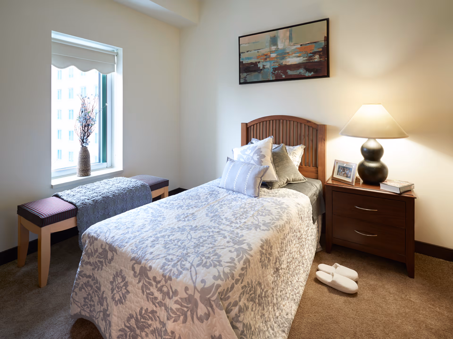 A cozy bedroom with a single bed covered in a white and gray floral patterned quilt and multiple pillows. Next to the bed is a wooden nightstand with a lamp, a framed photo, and a book. A pair of white slippers is placed on the carpeted floor beside the nightstand. A bench with a gray throw is positioned under a window with a vase of dried branches on the windowsill. A framed abstract painting hangs on the wall above the bed.