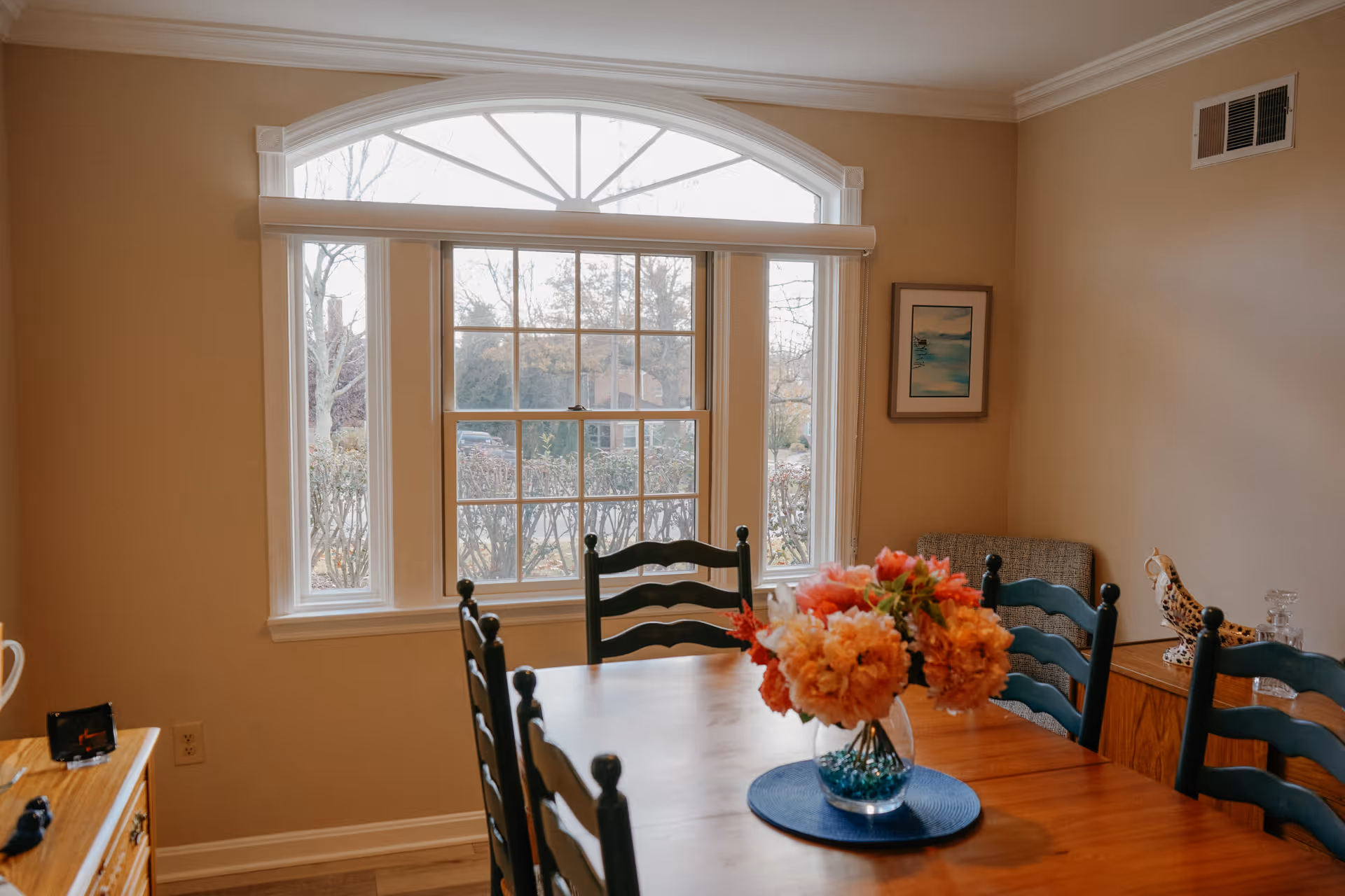 A dining room with a wooden table and six dark green chairs. A vase with orange and pink flowers sits on a blue placemat in the center of the table. There is a large window with white trim letting in natural light, a framed picture on the beige wall, and a wooden sideboard with decorative items.