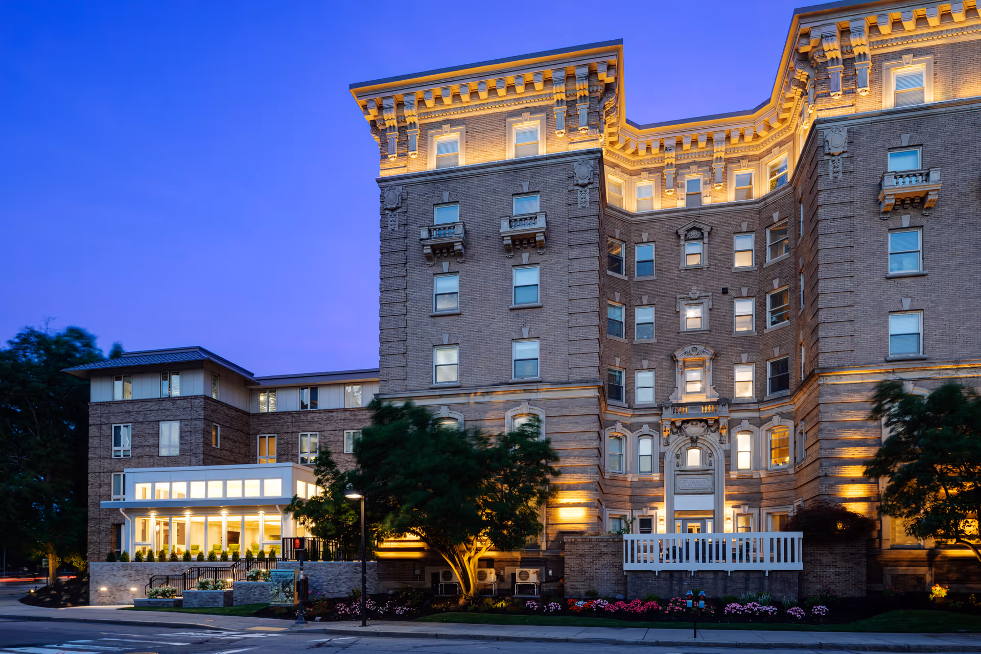 Exterior view of a large, multi-story brick building at dusk with warm lighting illuminating architectural details and windows. The building has a modern glass extension on the left side and landscaped greenery with flowers in front. The sky is clear and blue.