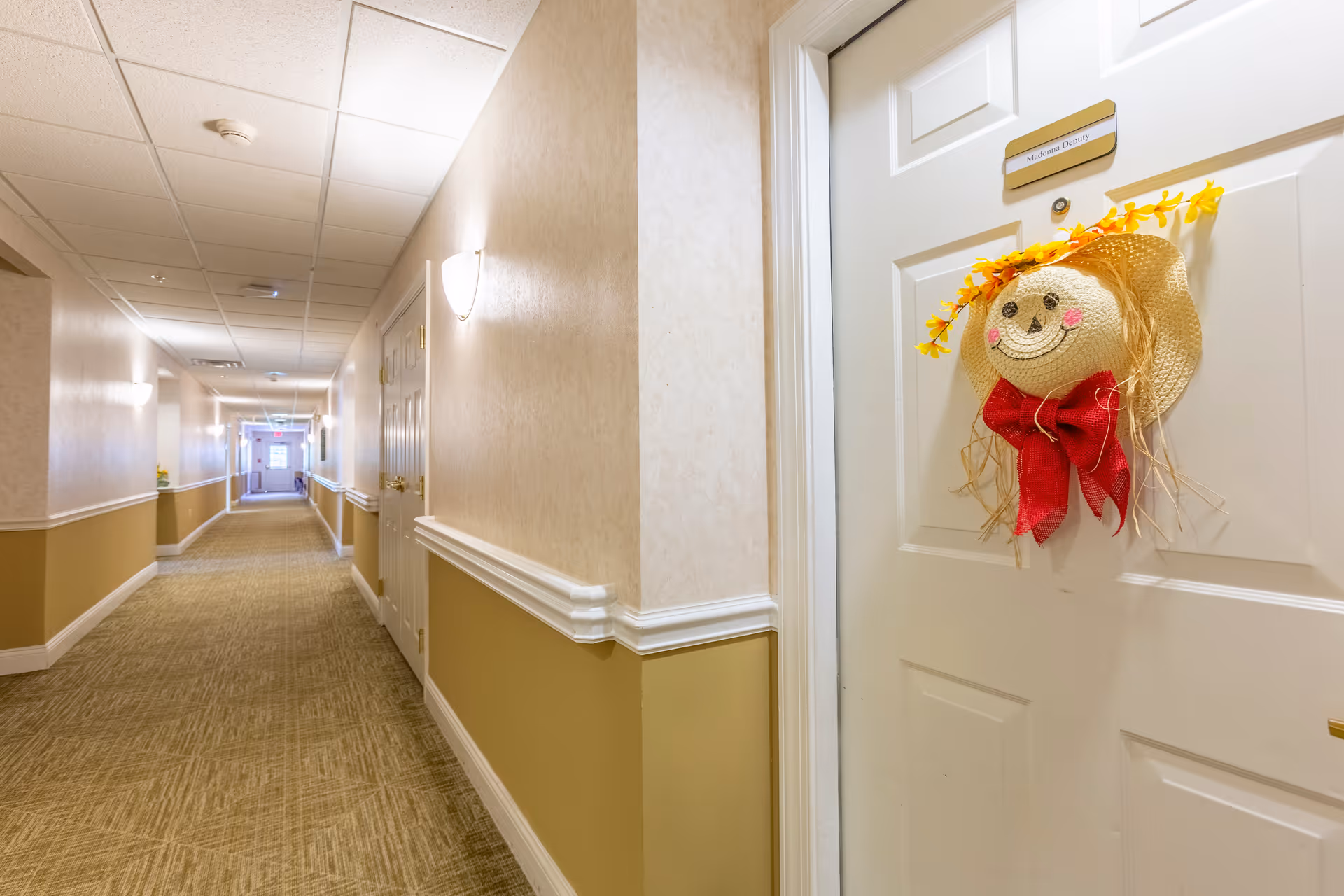 A long, well-lit hallway in a senior living facility with beige walls and carpeted floor. Several closed white doors line the hallway, one of which is decorated with a cheerful straw hat featuring a smiling face, red bow, and yellow flowers. The door has a nameplate above the decoration.