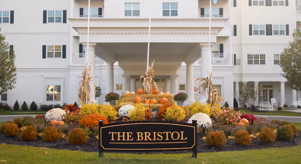 Front entrance of The Bristol Senior Living at Worthington Hills with a landscaped garden featuring pumpkins, hay bales, flowers, and cornstalks, in front of a large white building with columns and multiple windows.