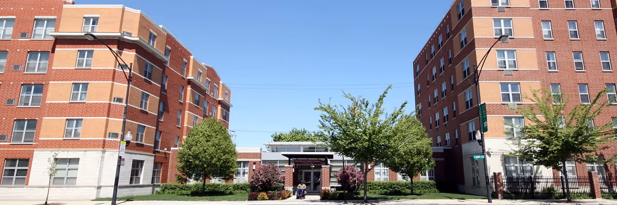 Front exterior view of Churchview Supportive Living facility showing two large brick buildings on either side with multiple windows, a central entrance with a covered walkway, trees, shrubs, and a clear blue sky.