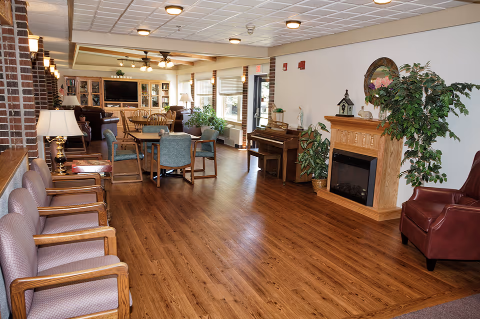 A spacious and well-lit common area in a retirement community featuring wooden flooring, multiple seating arrangements including armchairs and dining tables with chairs, a piano, a fireplace with decorative plants, and a large TV with a wooden cabinet in the background.
