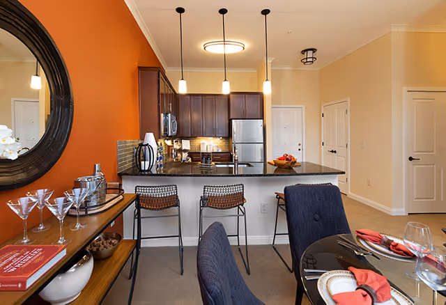 Interior view of a senior living facility apartment featuring a kitchen with dark wood cabinets, stainless steel appliances, and a black countertop island with three bar stools. Adjacent to the kitchen is a dining area with a glass table set with plates, napkins, and glasses. A console table with decorative items and a round mirror is visible on the left side. The walls are painted in warm tones, including an orange accent wall.