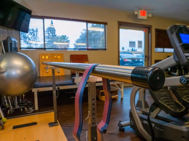 Interior view of a rehabilitation or fitness room with exercise equipment including a balance ball, parallel bars with resistance bands, and a treadmill. There is a window with blinds and an exit door showing parked cars outside.