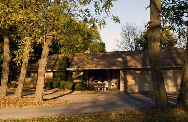 Single-story brick building with a sloped roof surrounded by trees and bushes. There is a driveway in front and a covered patio area with a table and chairs. The scene is lit by late afternoon sunlight with some shadows from the trees.