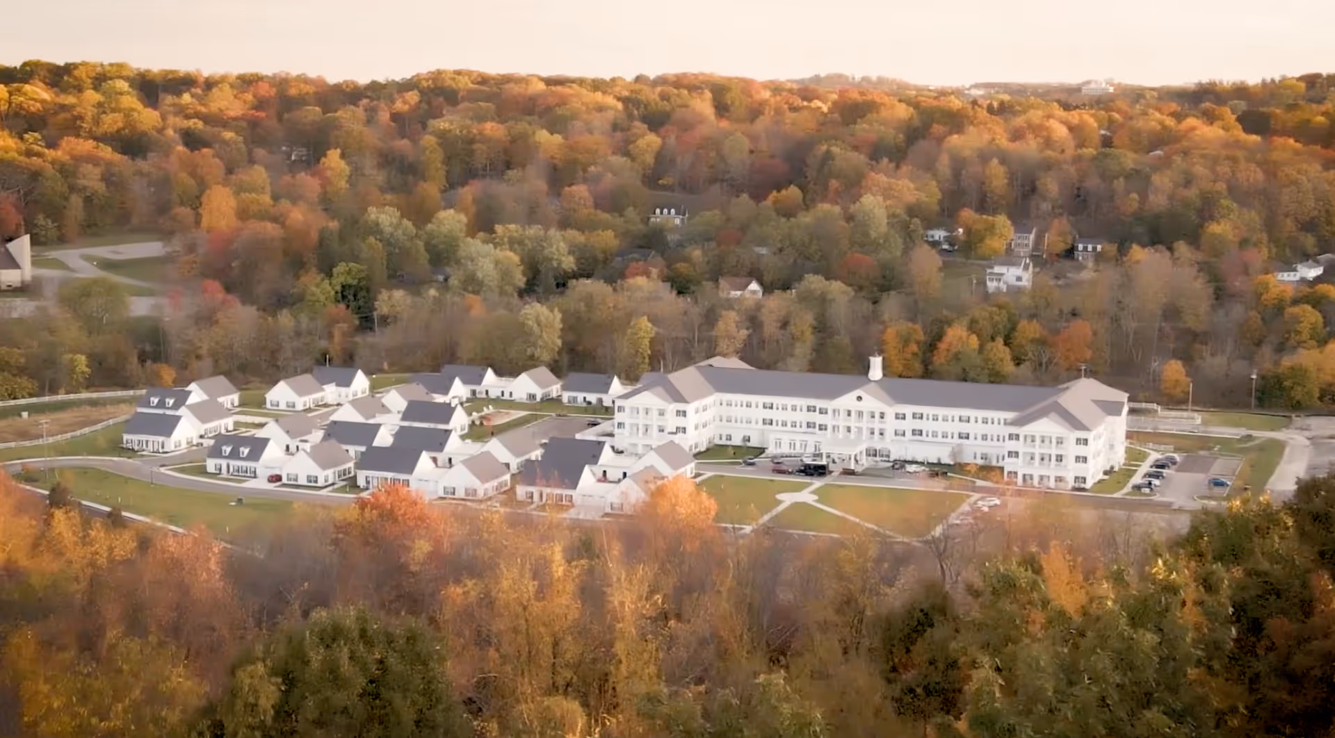 Aerial view of Ashton Commons senior living facility surrounded by trees with autumn foliage. The facility includes a large main building and several smaller white residential cottages arranged in a campus-like layout with parking areas and green lawns.