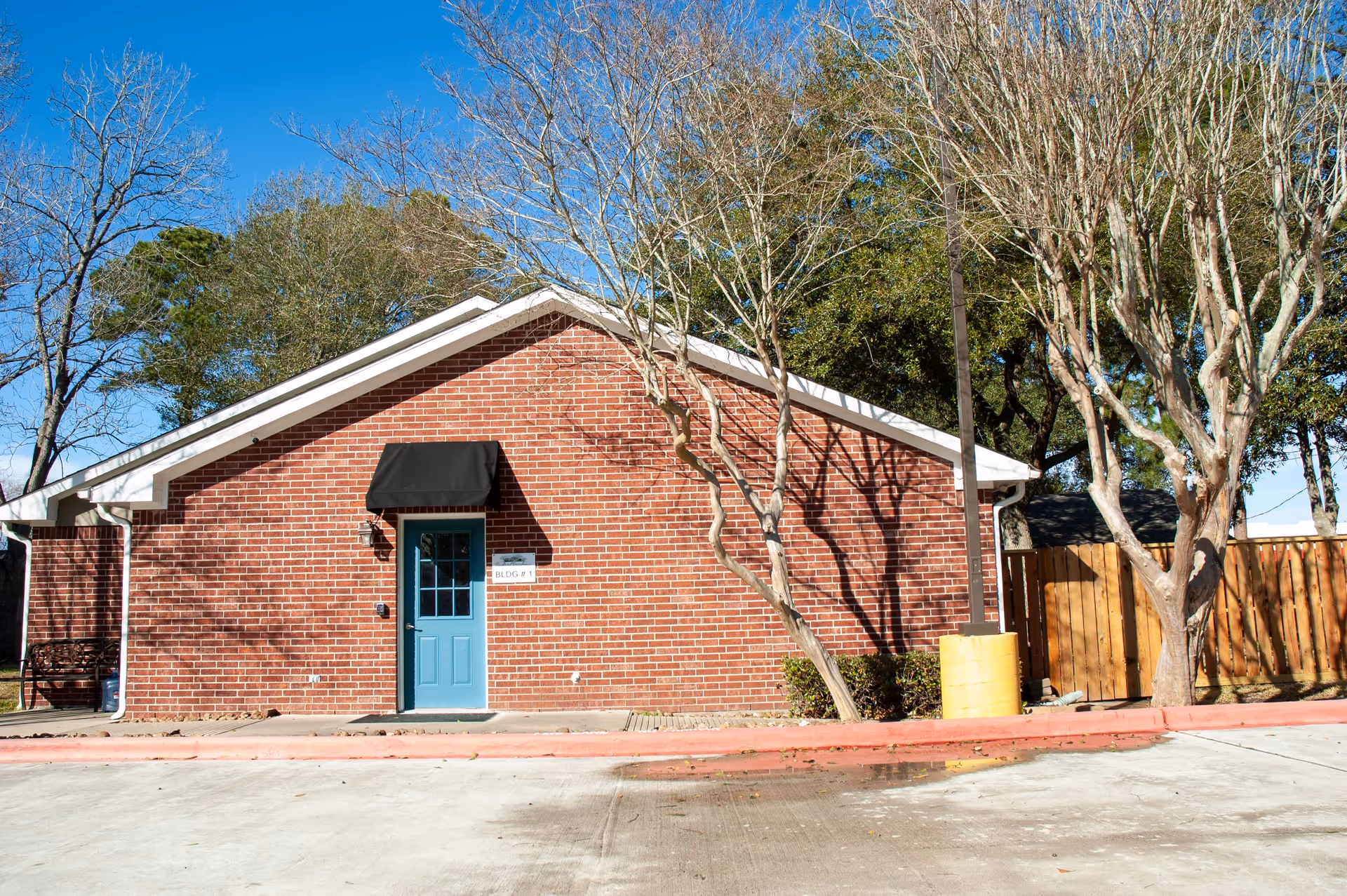 Exterior view of a single-story brick building with a blue door under a black awning, surrounded by leafless trees and a wooden fence, with a clear blue sky in the background.