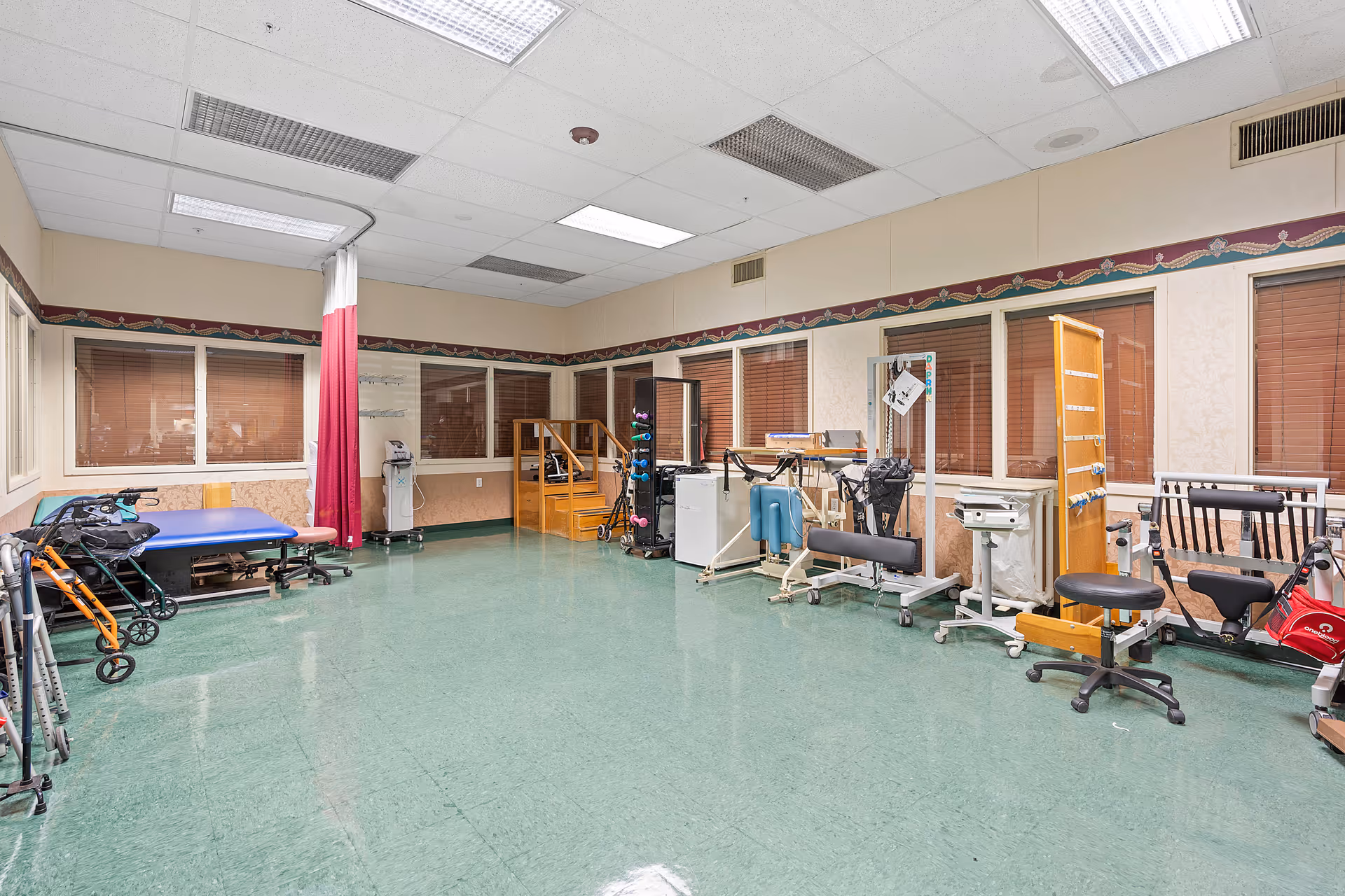 A spacious rehabilitation or physical therapy room with green flooring, various exercise and therapy equipment including walkers, a treatment table, weights, and parallel bars. The room has beige walls with a decorative border near the ceiling and several windows with closed blinds.