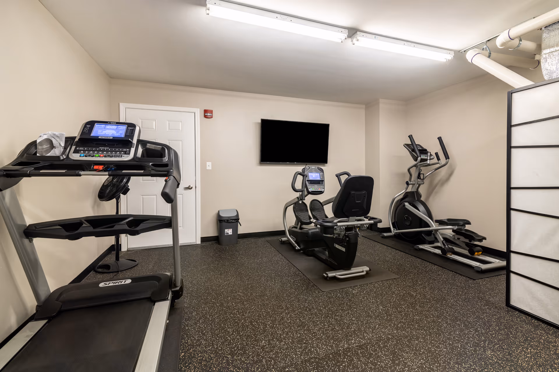 A small fitness room with a treadmill, a recumbent exercise bike, and an elliptical machine. The room has beige walls, a black speckled rubber floor, a wall-mounted flat screen TV, a trash can, and a white door. There is also a black standing fan and a partial view of a room divider on the right.