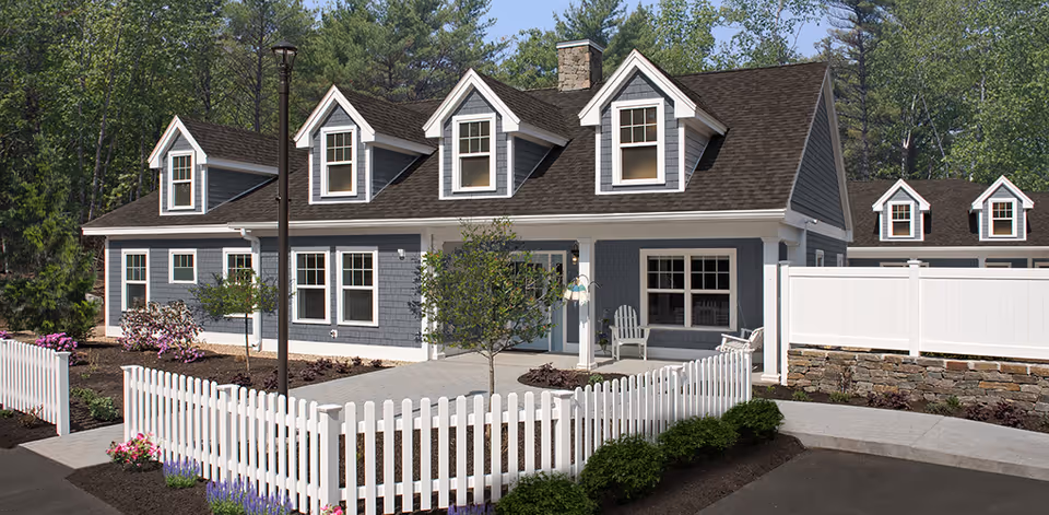 Exterior view of a charming residential building with gray siding, white trim, and multiple dormer windows on the roof. The building is surrounded by a white picket fence, small trees, and landscaped flower beds. There is a paved walkway and a lamp post in front of the building, with a wooded area in the background.