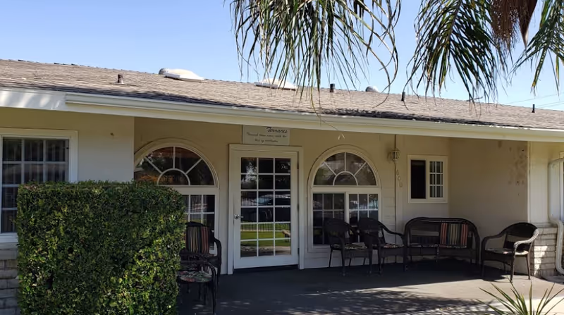 Exterior view of a single-story building with a covered patio area featuring several wicker chairs and a bench. The building has arched windows and a glass door with a sign above it. There are palm tree leaves hanging down from the top of the image and a green hedge on the left side.