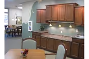 Interior view of a senior living facility showing a kitchenette area with wooden cabinets, a countertop, and under-cabinet lighting. In the foreground, there is a table with chairs, and in the background, a dining area with a large table and chairs is visible. The walls are painted in soft colors with a decorative design.