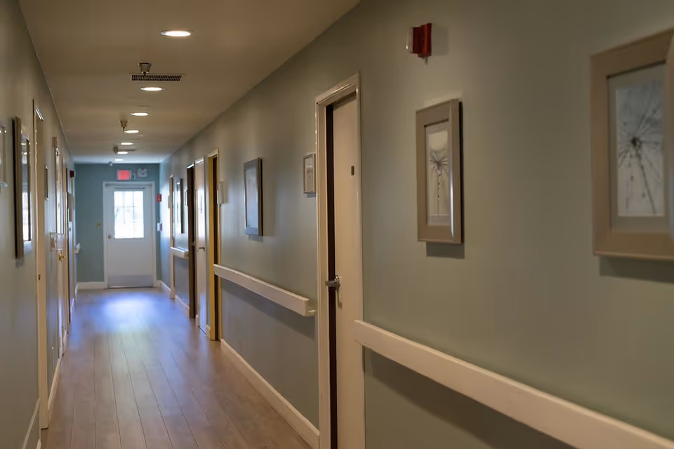 A well-lit hallway in a senior living facility with light green walls, wooden flooring, and several closed doors on both sides. The hallway is decorated with framed pictures and has handrails along the walls. A door with a window is visible at the far end of the hallway.