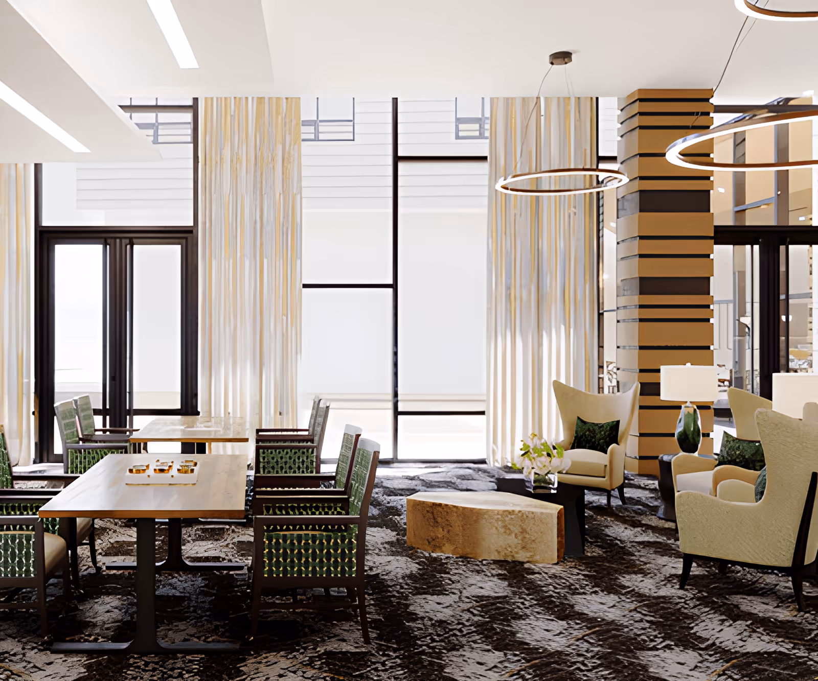 A modern senior living facility common area with wooden tables and green cushioned chairs on the left, and beige armchairs with green pillows arranged around a stone coffee table on the right. The room features large windows with sheer curtains, a patterned carpet, and circular pendant lights hanging from the ceiling.