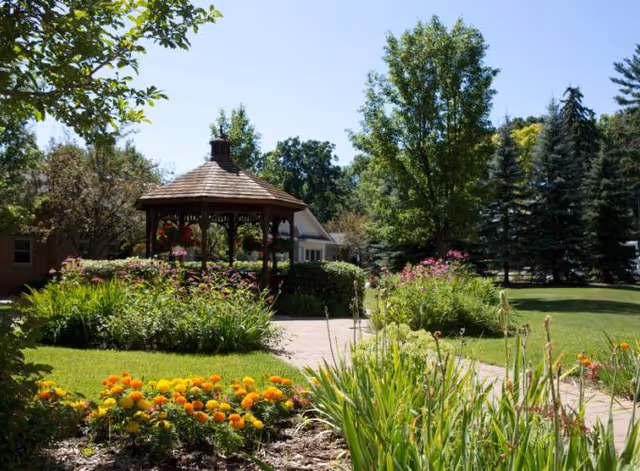 A sunny outdoor garden area with a wooden gazebo surrounded by lush greenery, colorful flowers, and tall trees. A paved walkway leads through the garden, and a building is partially visible in the background.