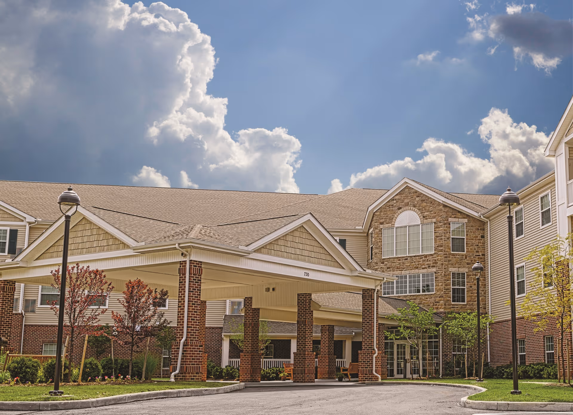 Exterior view of Danbury Westerville senior living facility showing a large covered entrance with brick pillars, well-maintained landscaping, and a partly cloudy sky above.
