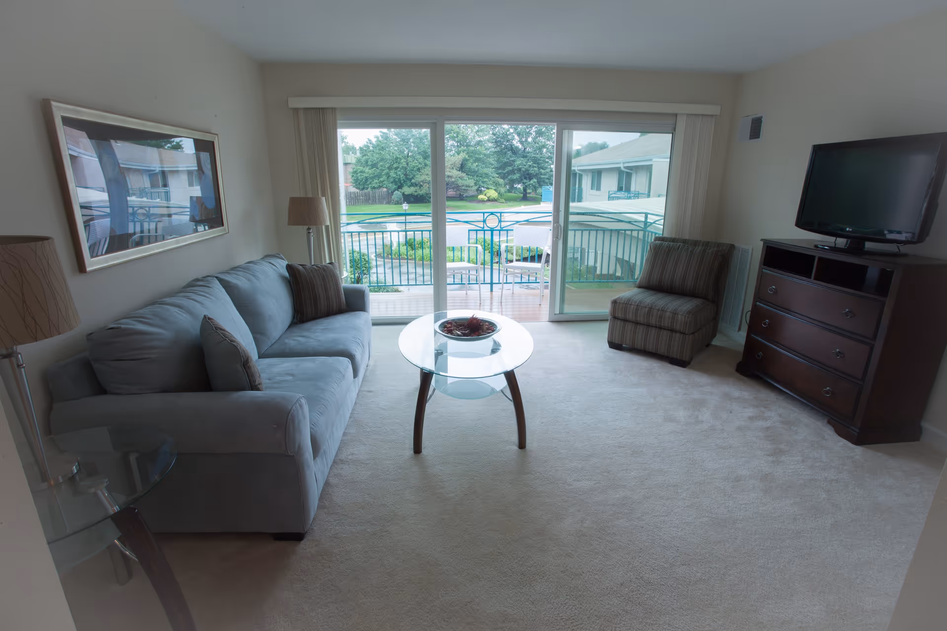 A living room with a light gray sofa, a striped armchair, a glass coffee table with a decorative bowl, a wooden dresser with a flat-screen TV on top, and a floor lamp. The room has sliding glass doors that open to a balcony overlooking a pool and greenery outside.