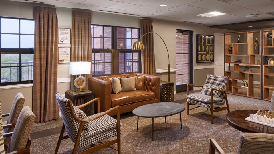 A cozy senior living common area with a brown leather sofa, two patterned armchairs, a round coffee table, and a floor lamp. The room has large windows with brown plaid curtains, a wooden shelving unit with decorative items, and a chess set on a round wooden table. The walls are light-colored with framed artwork and a door leading outside.