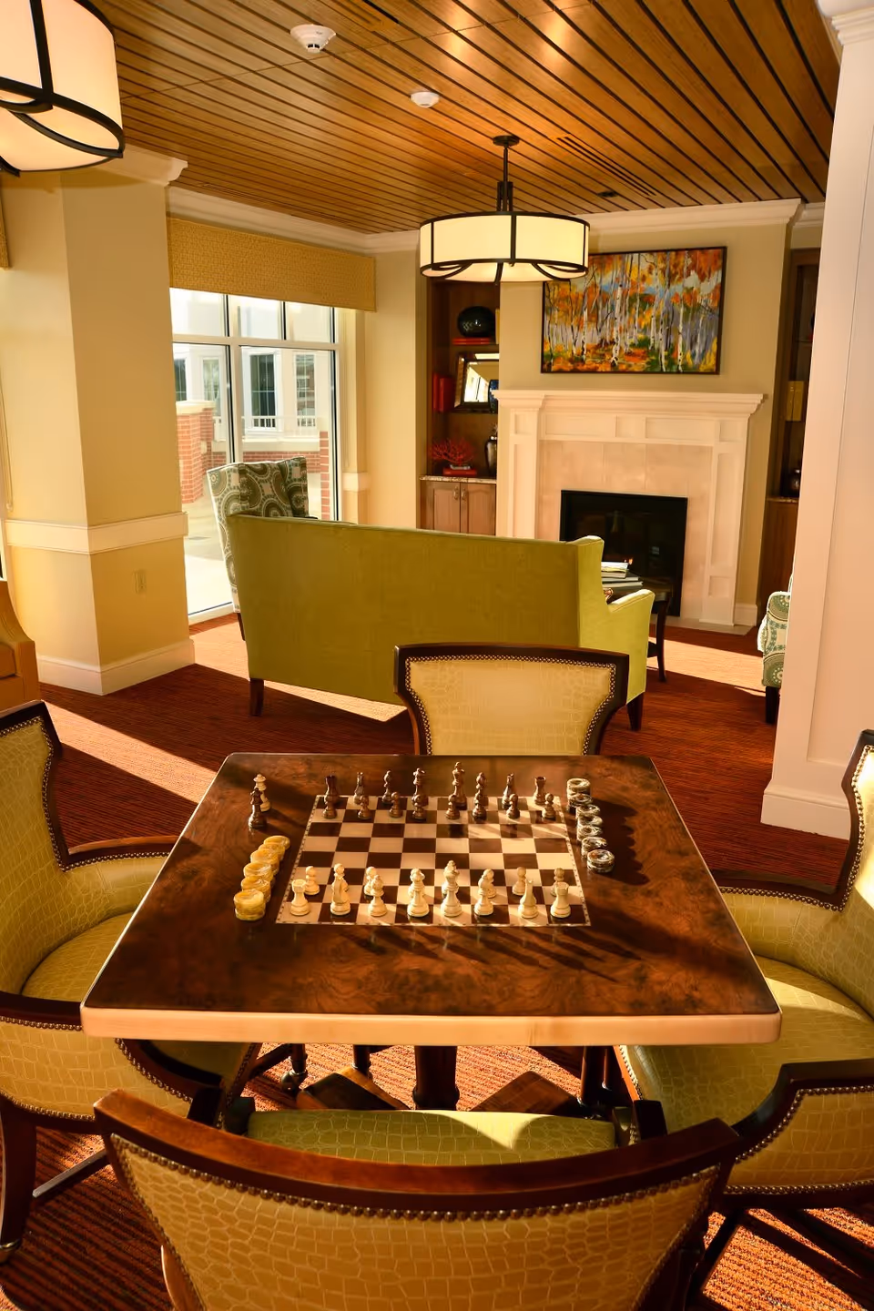 A cozy living room area with a wooden chess table in the foreground surrounded by four green upholstered chairs. Behind the table, there is a green sofa facing a white fireplace with a colorful painting above it. The room has large windows letting in natural light, a wooden slatted ceiling, and warm-toned carpeting.